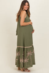 woman wearing an olive green sleeveless maternity maxi dress with floral embroidery and holding her baby bump
