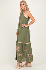 woman wearing an olive green maxi dress with a halter neckline and embroidered palm trees at the hem