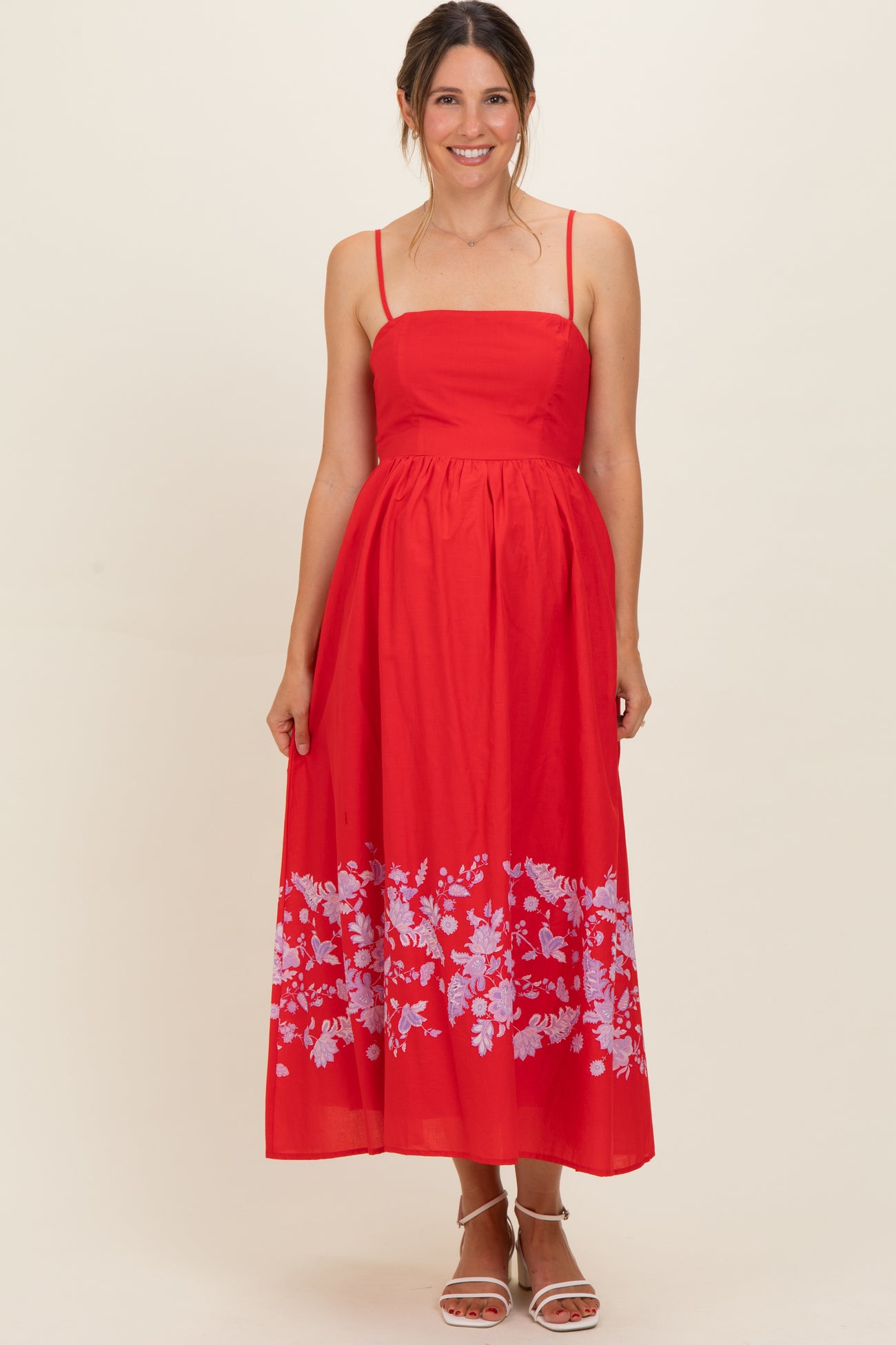 woman wearing a red maternity maxi dress with floral print at the hem and spaghetti straps, showing her baby bump