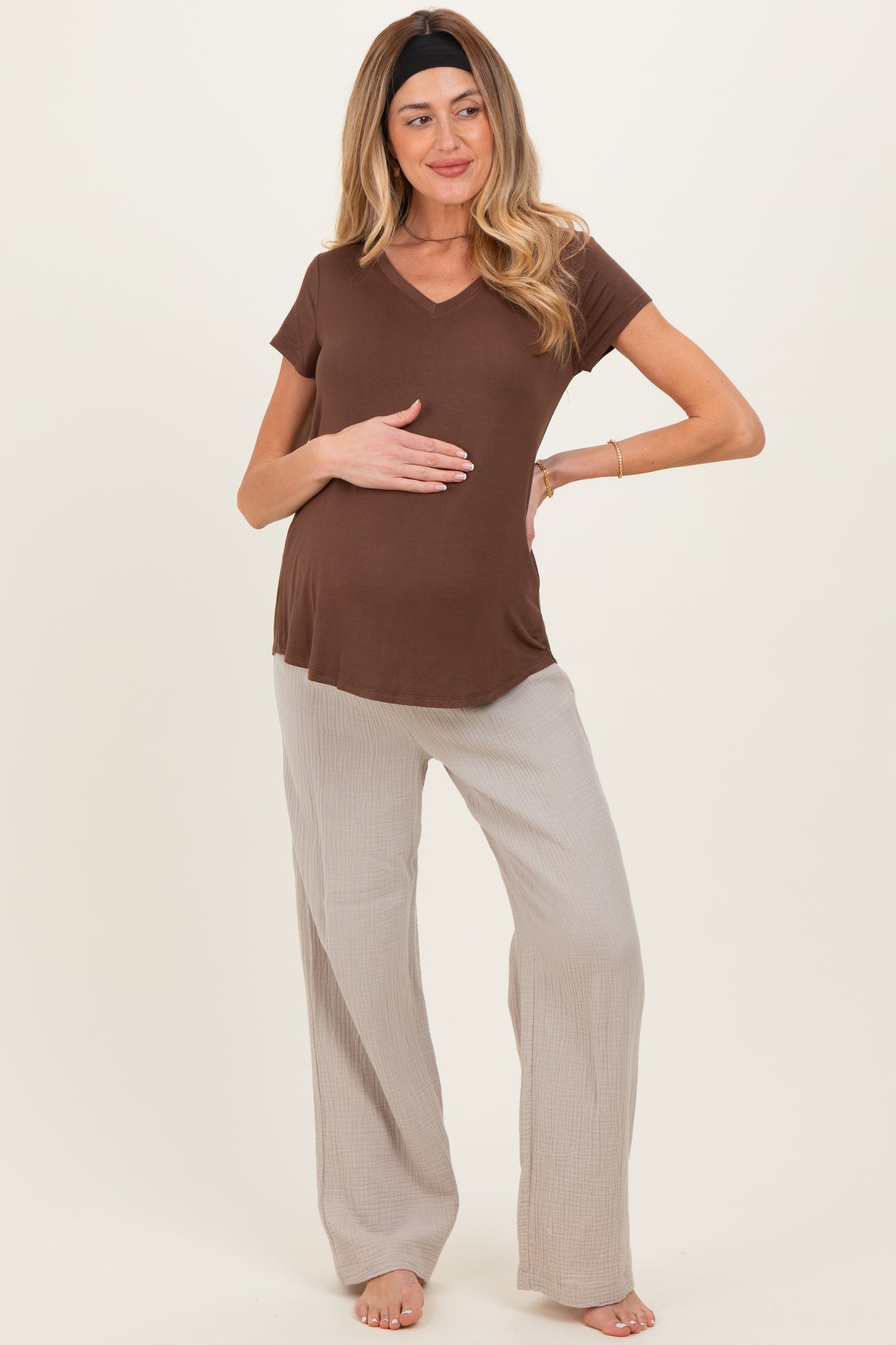 woman wearing a brown short sleeve maternity top and light beige wide-leg pants, holding her baby bump