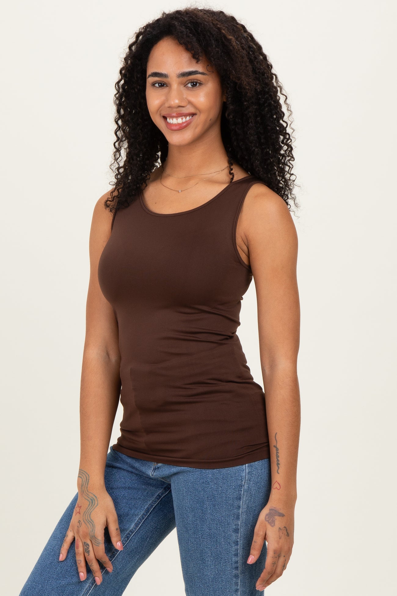 woman wearing a brown fitted tank top and blue jeans, smiling with curly hair