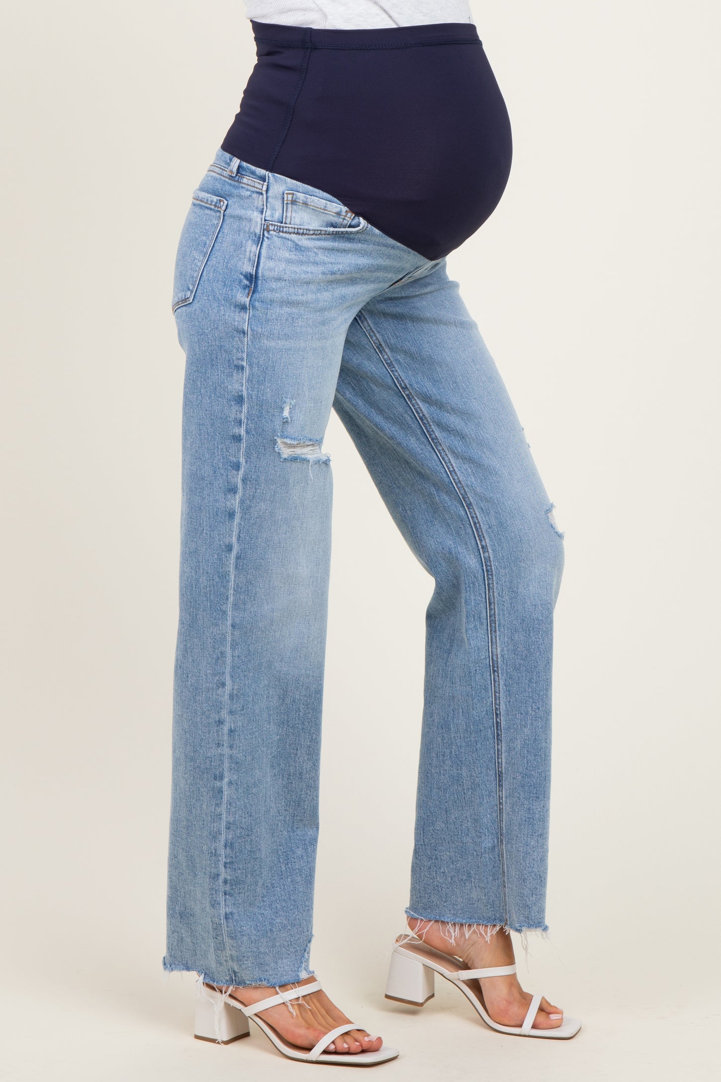 woman wearing light blue distressed maternity jeans with a navy belly panel and white block-heeled sandals, baby bump visible