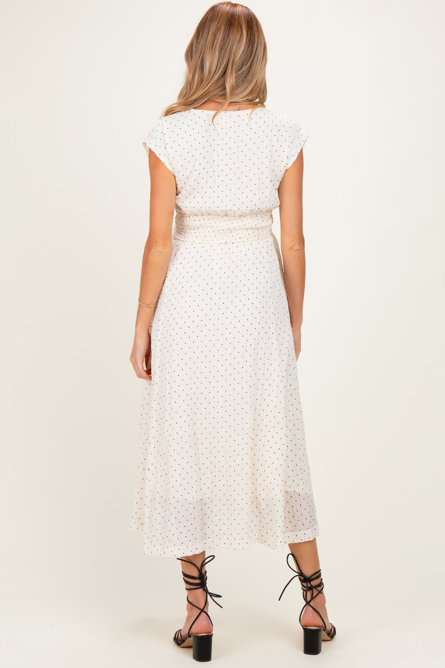 woman wearing a cream polka dot maternity dress with cap sleeves and a tie waist, viewed from the back