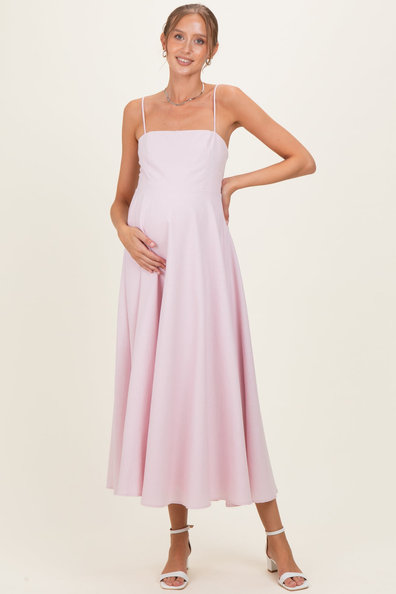 woman wearing a light pink spaghetti strap maternity dress and gently holding her baby bump