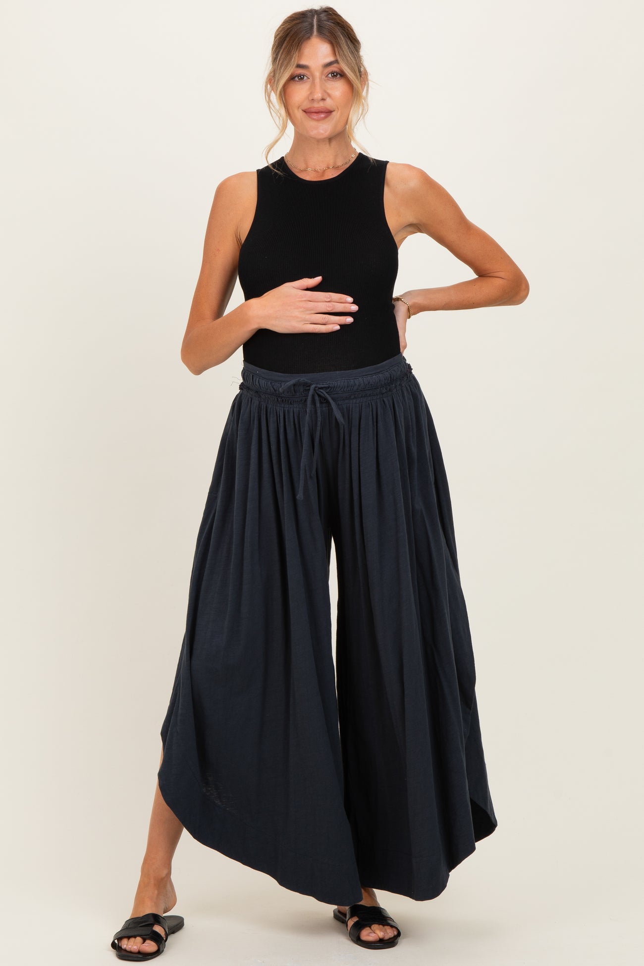 woman wearing a black sleeveless maternity top and dark blue wide-leg maternity pants, holding her baby bump