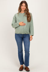 woman wearing a green maternity cardigan and blue jeans, gently cradling her baby bump
