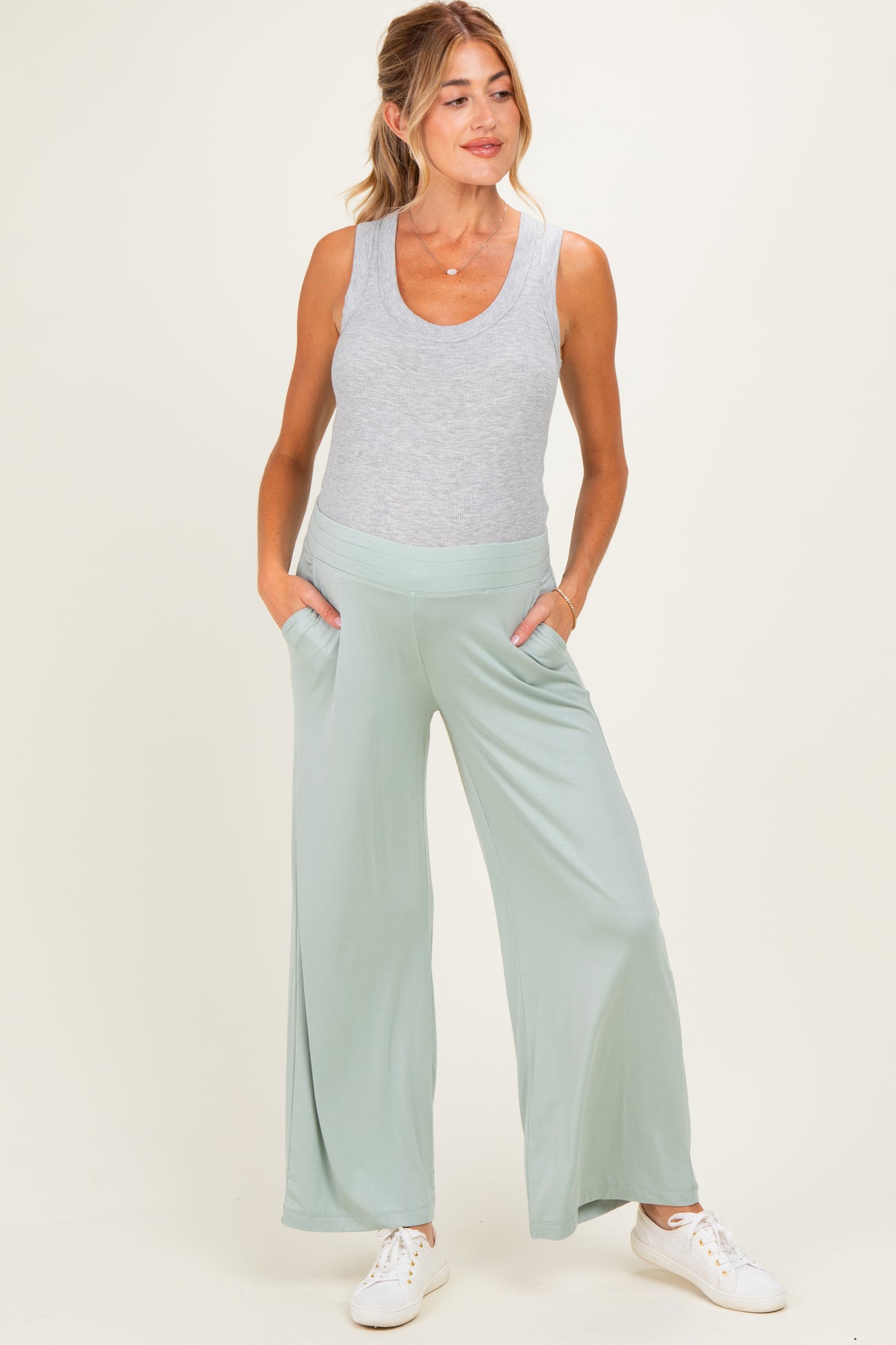 woman wearing a light grey tank top and light green wide-leg maternity pants with pockets and a visible baby bump