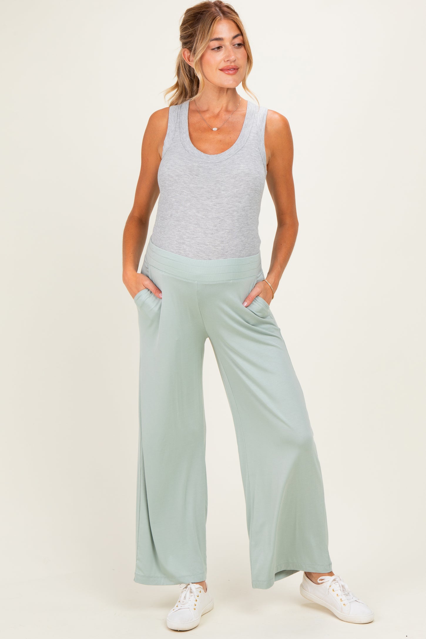 woman wearing a light grey tank top and light green wide-leg maternity pants with pockets and a visible baby bump