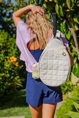 woman wearing a light purple short sleeve top and navy shorts, carrying a white quilted backpack while walking outdoors