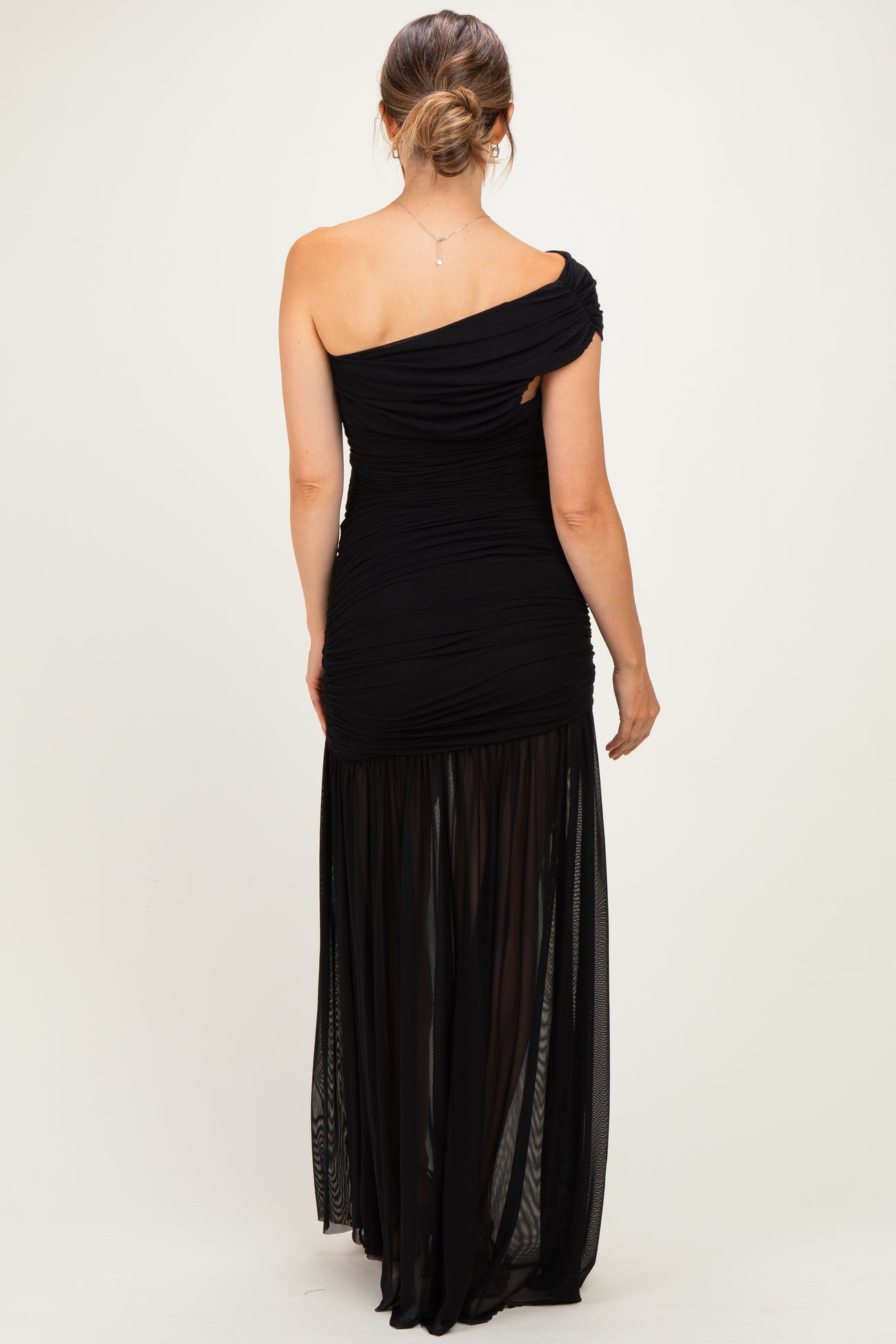 woman wearing a black one-shoulder maternity dress with a fitted bodice and flowing skirt, showing off a baby bump from the back