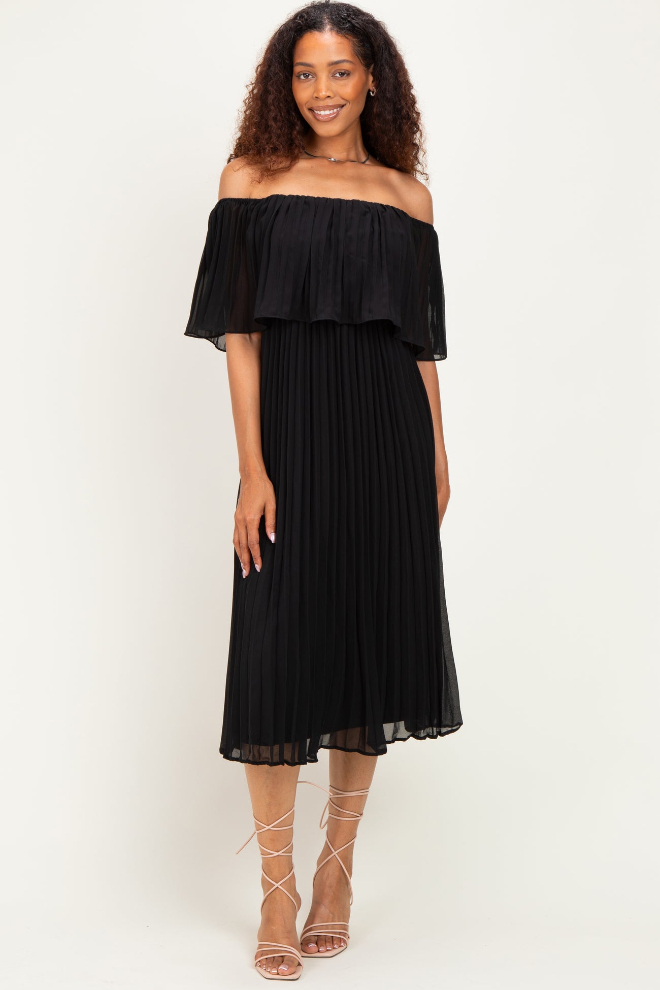 woman wearing a black off-the-shoulder pleated dress with a flowing ruffle detail and strappy sandals