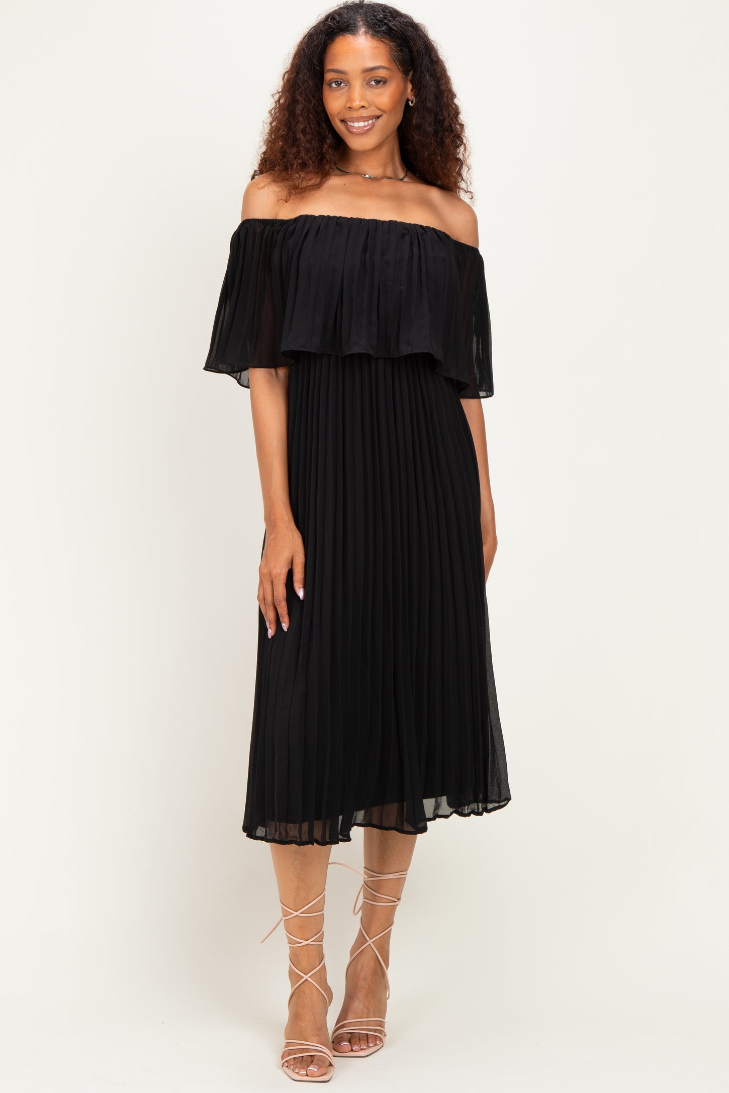 woman wearing a black off-the-shoulder pleated dress with a flowing ruffle detail and strappy sandals