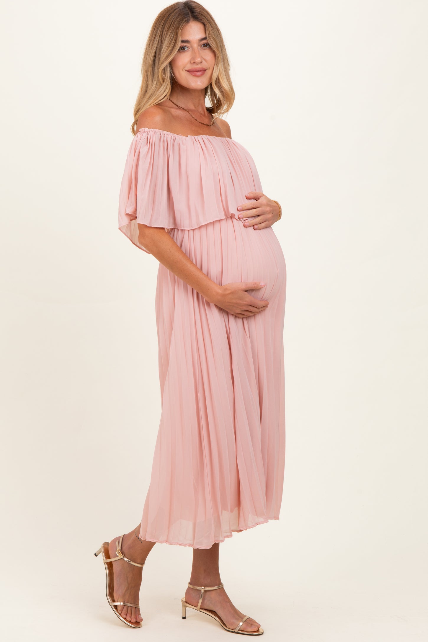 woman wearing a light pink off-the-shoulder maternity dress and holding her baby bump