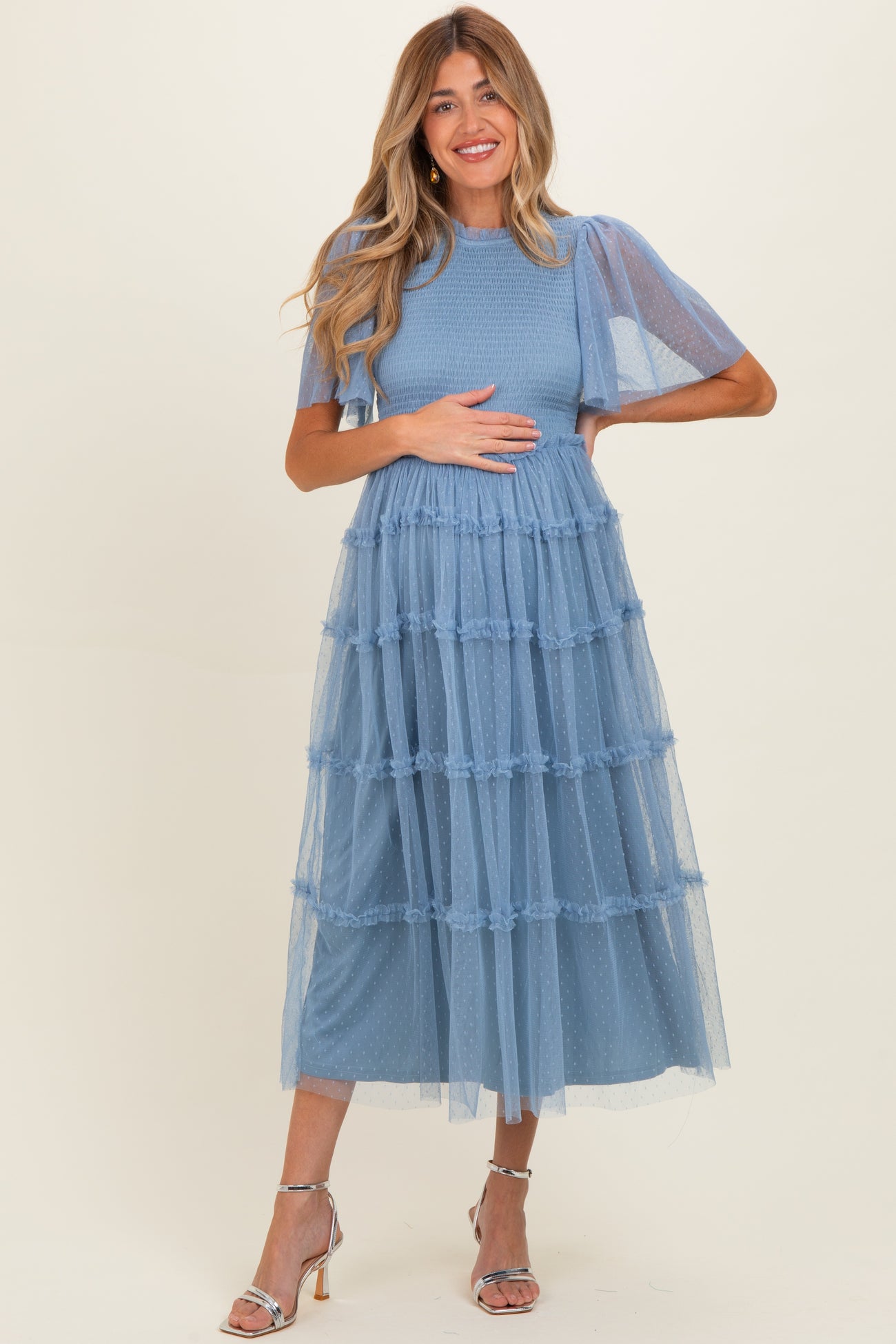 woman wearing a light blue maternity dress with tiered tulle layers and short sleeves, holding her baby bump