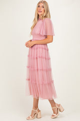 woman wearing a pink tiered tulle dress with short sleeves and ruffle details, standing sideways on a neutral background