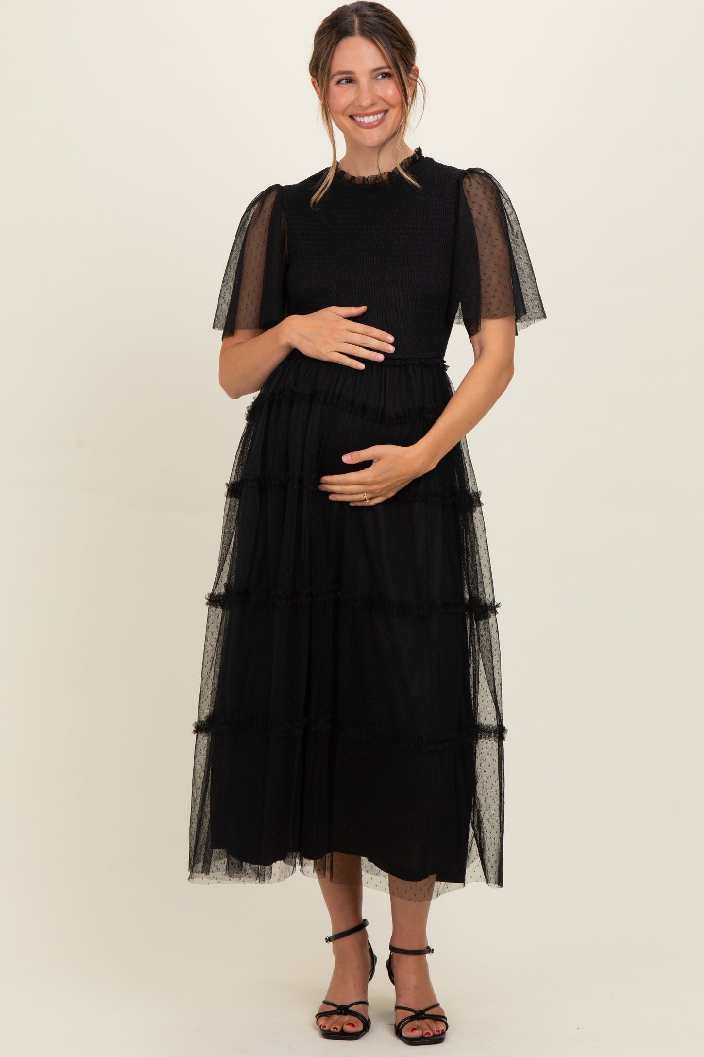 woman wearing a black maternity dress with layered tulle skirt and sheer puff sleeves, holding her baby bump