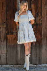 woman wearing a blue and white striped smocked dress with short sleeves and lace trim, standing in front of a wooden backdrop
