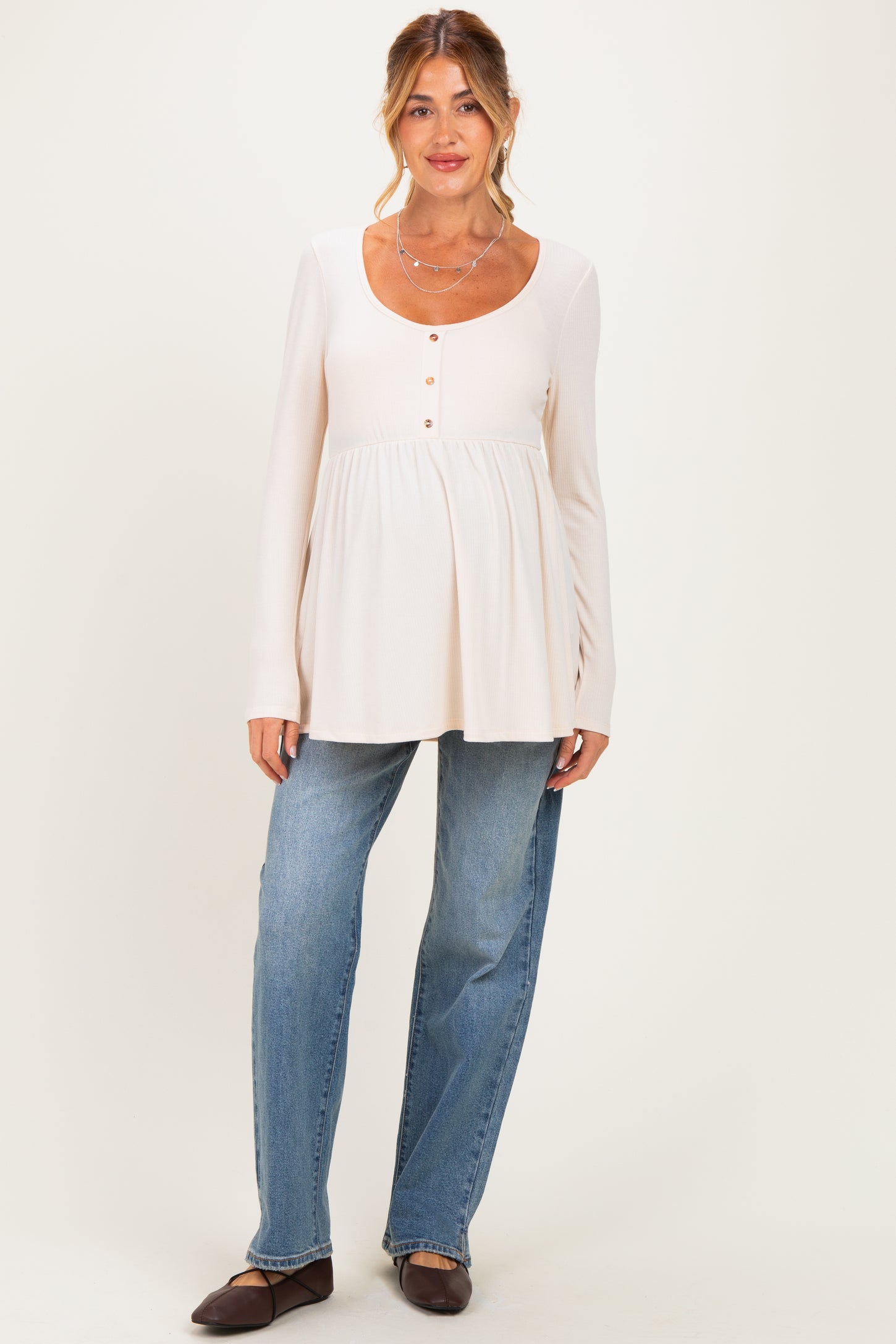 woman wearing a light beige long sleeve maternity top with button details and a baby bump, paired with blue jeans