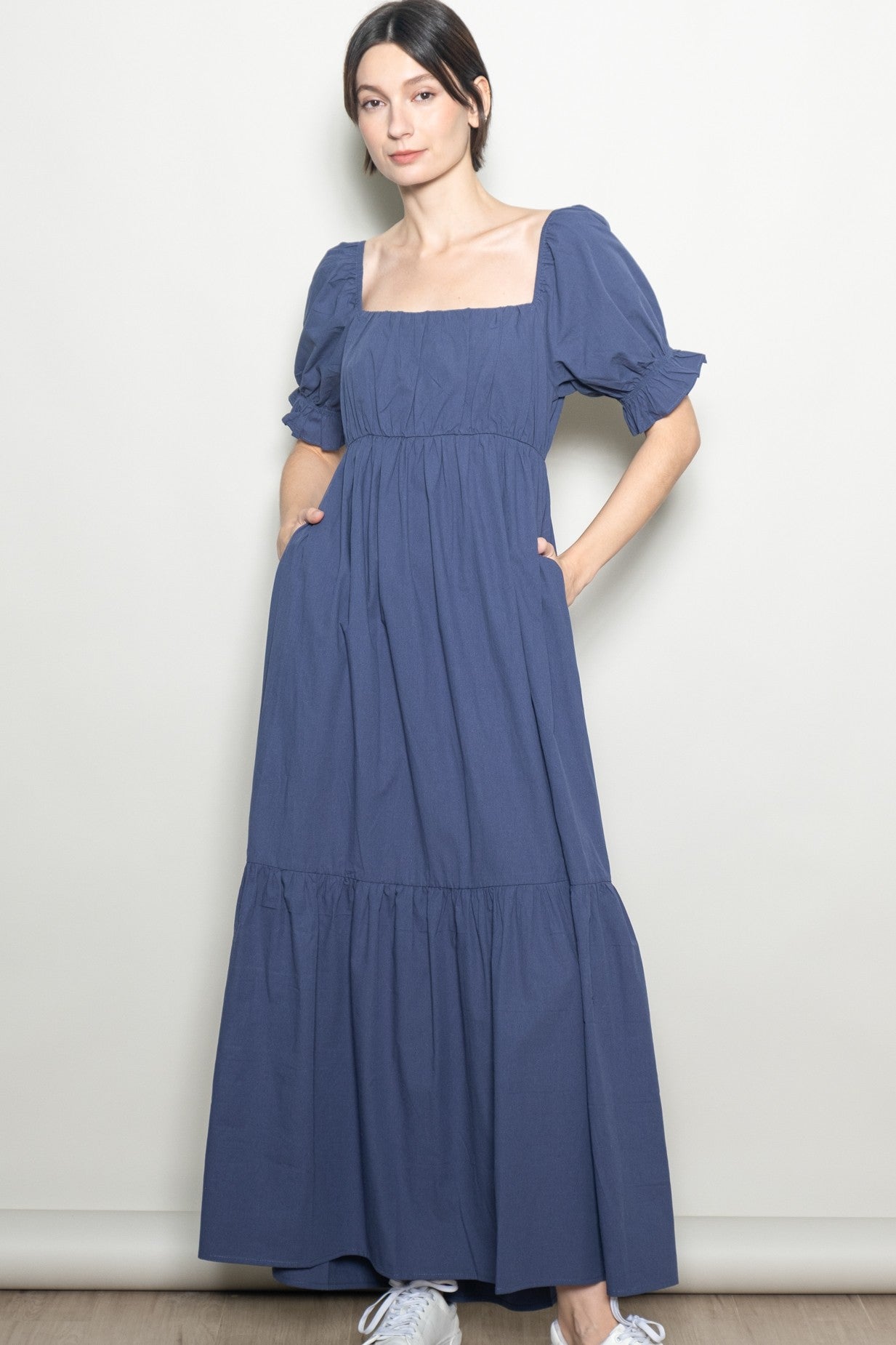 woman wearing a navy blue maxi dress with square neckline and puff sleeves, standing with hands in pockets