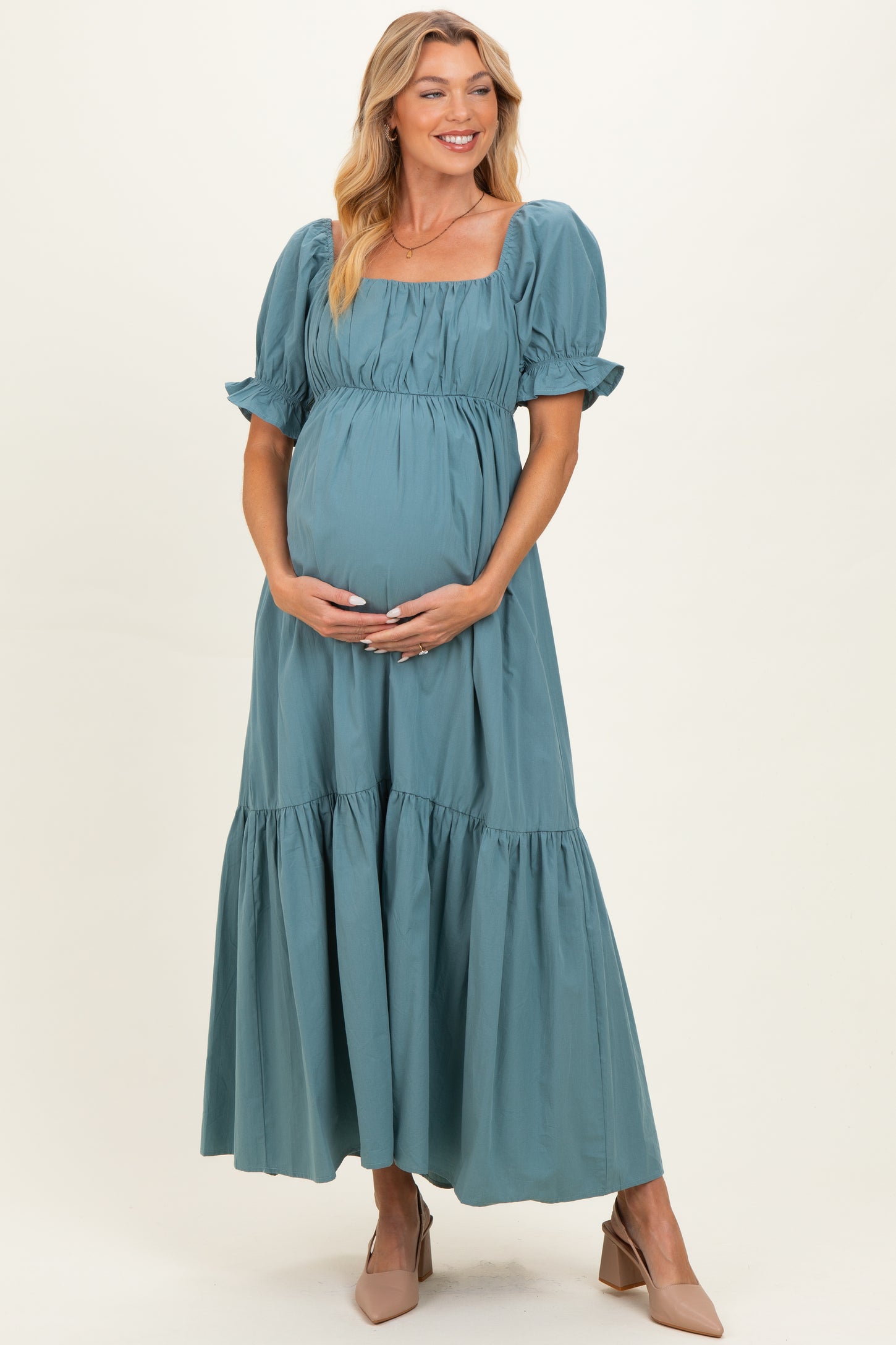 woman wearing a teal maternity maxi dress with puff sleeves and a tiered skirt, holding her baby bump