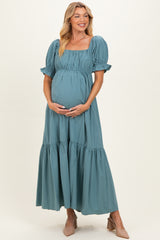 woman wearing a teal maternity maxi dress with puff sleeves and a tiered skirt, holding her baby bump
