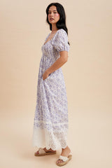 woman wearing a lavender floral maxi dress with puff sleeves and lace trim, standing with hands in pockets