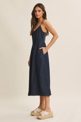 woman wearing a dark denim midi dress with adjustable straps and side pockets, paired with beige wedge sandals