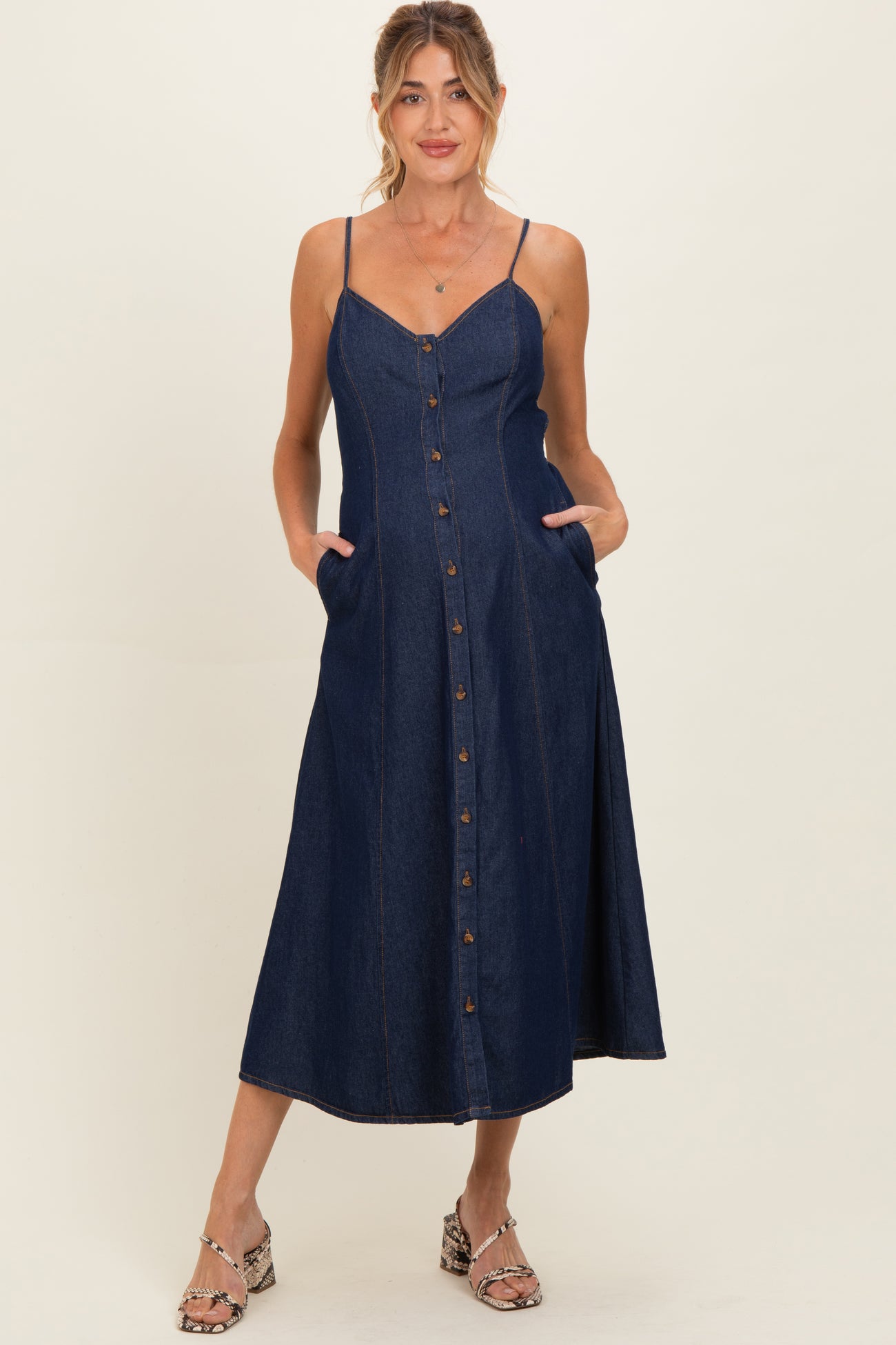 woman wearing a dark denim maternity dress with button-down front and pockets, featuring thin straps and a flowy skirt