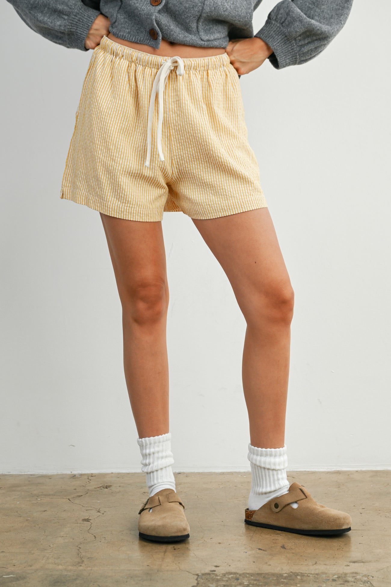 woman wearing yellow and white striped shorts with an elastic waistband and gray sweater, paired with beige clogs and white socks