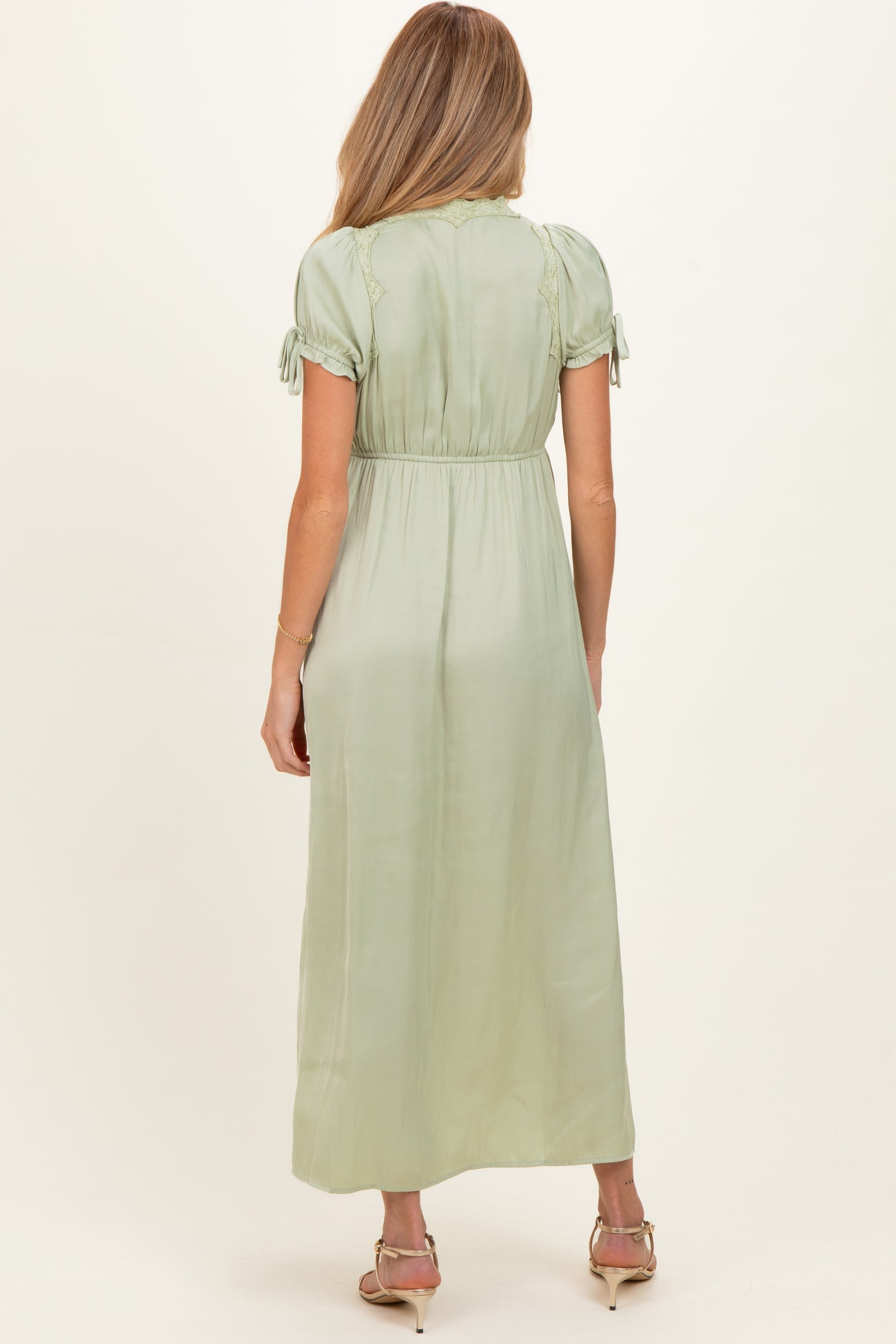 woman wearing a sage green short sleeve maternity maxi dress with tie details and a flowy silhouette from the back