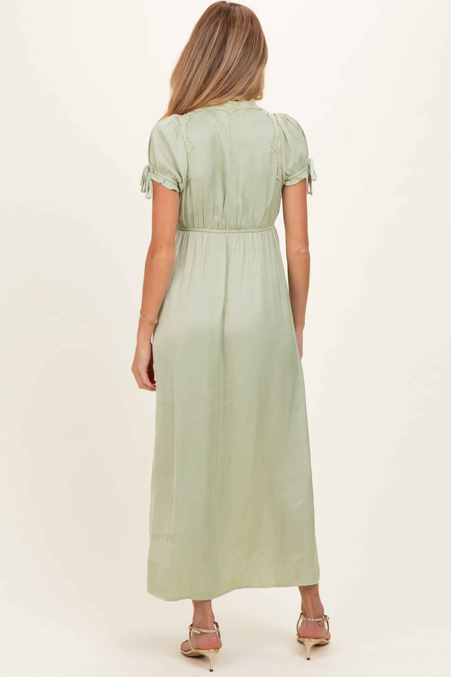 woman wearing a sage green short sleeve maternity maxi dress with tie details and a flowy silhouette from the back
