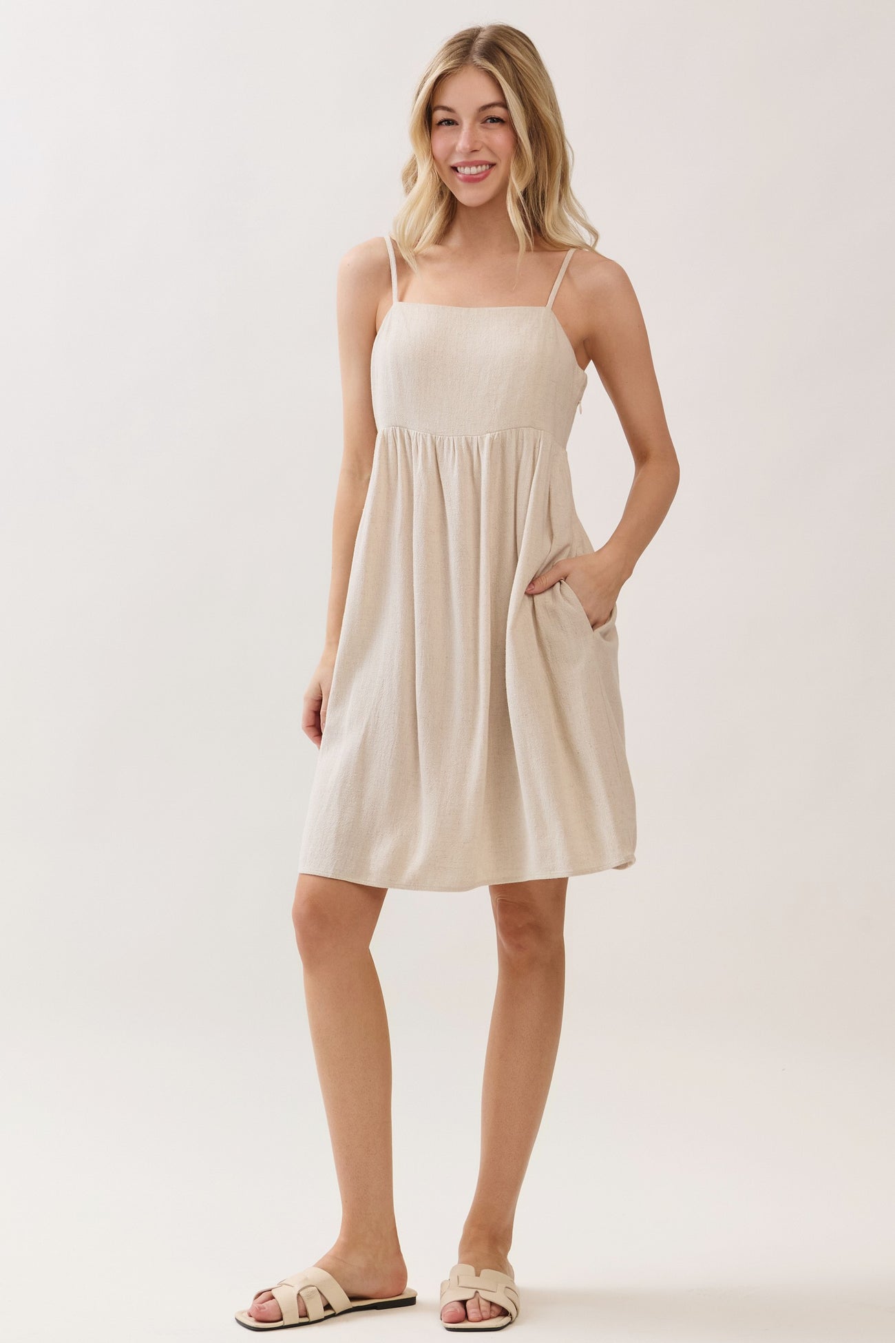 woman wearing a beige sleeveless sundress with a gathered waist and pockets, standing against a light background