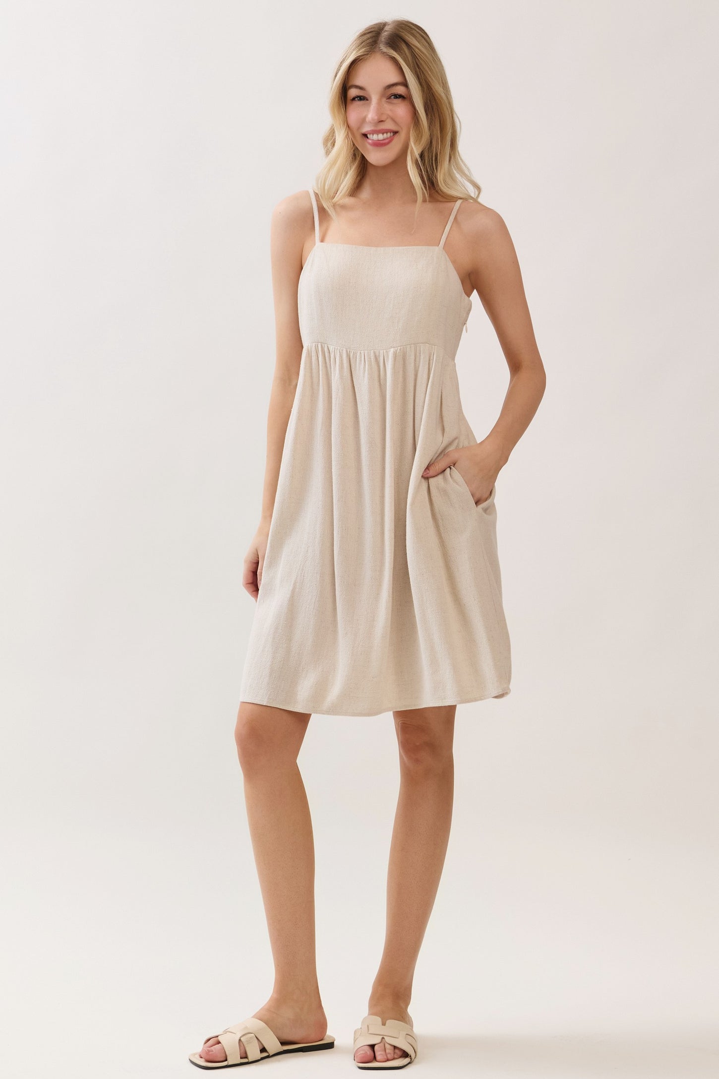 woman wearing a beige sleeveless sundress with a gathered waist and pockets, standing against a light background