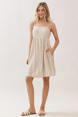 woman wearing a beige sleeveless sundress with a gathered waist and pockets, standing against a light background