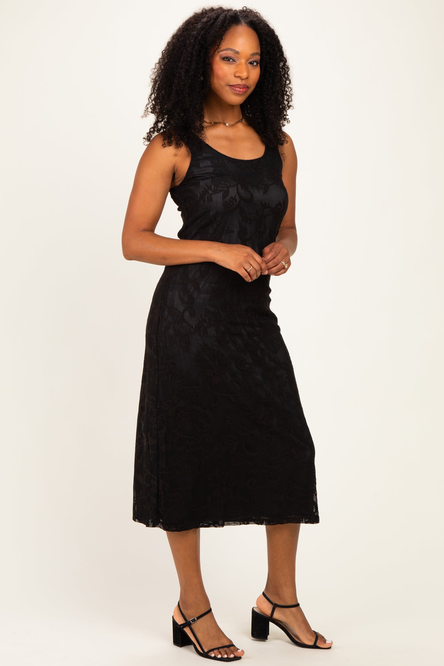 woman wearing a black sleeveless midi dress with a floral lace pattern and paired with black heeled sandals