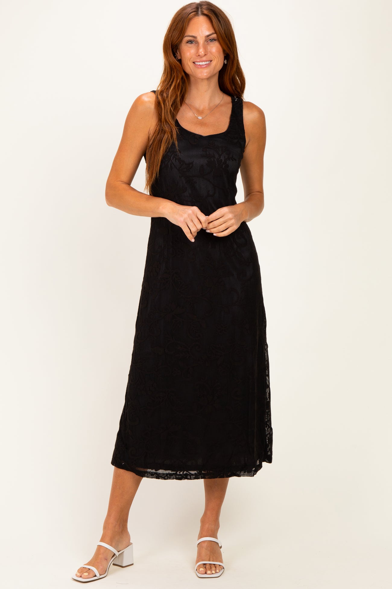 woman wearing a black sleeveless midi dress with a floral lace overlay and standing on a light background