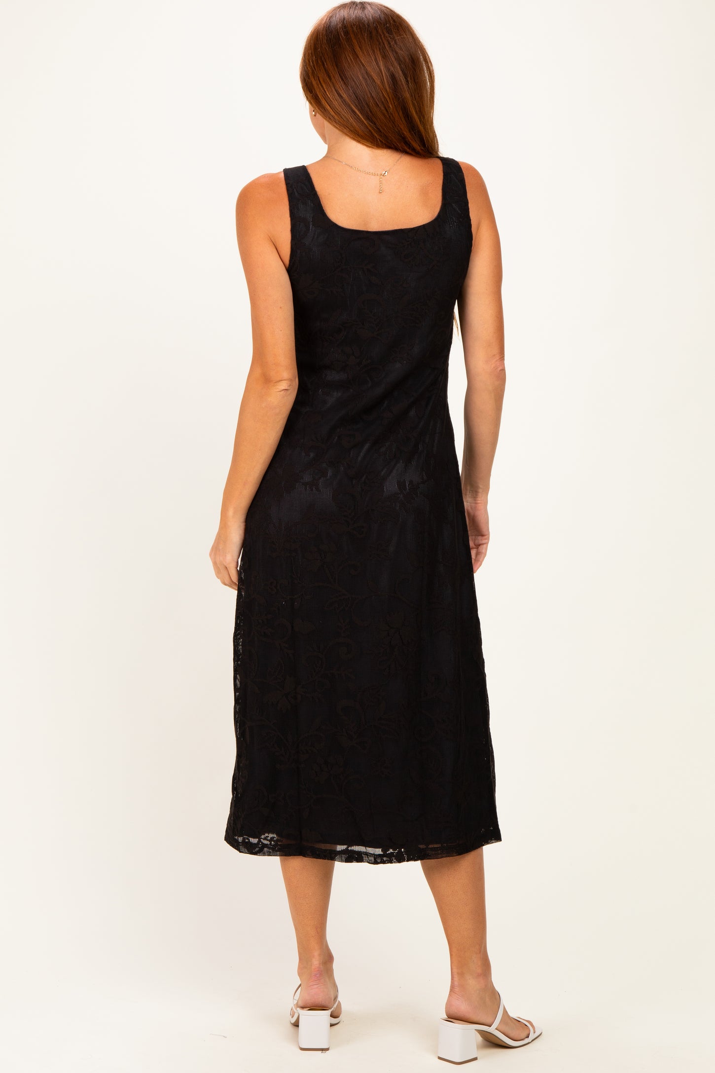 woman wearing a black sleeveless midi dress with a floral lace design, standing with her back to the camera