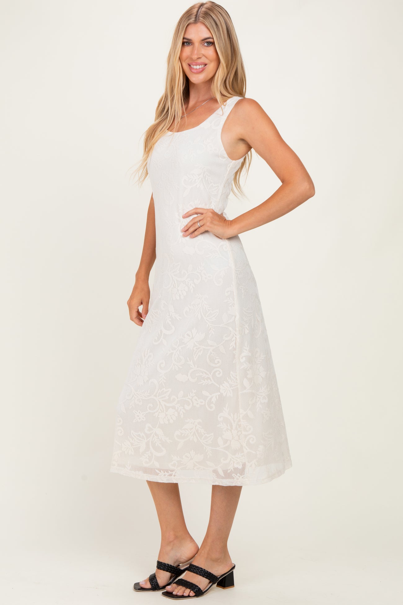 woman wearing a white midi dress with floral embroidery and adjustable straps, standing sideways with a smile
