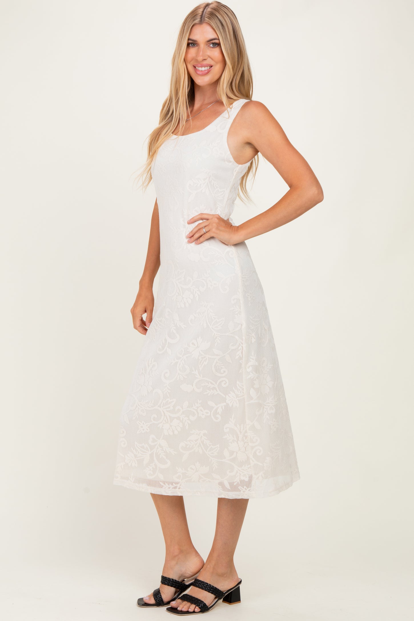 woman wearing a white midi dress with floral embroidery and adjustable straps, standing sideways with a smile