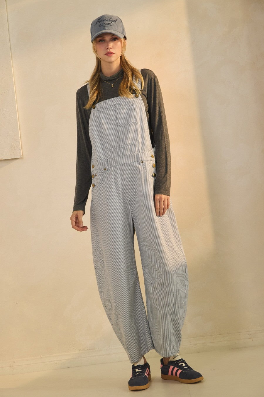 woman wearing blue and white striped overalls with side buttons and a long-sleeve gray top, paired with a gray cap and sneakers