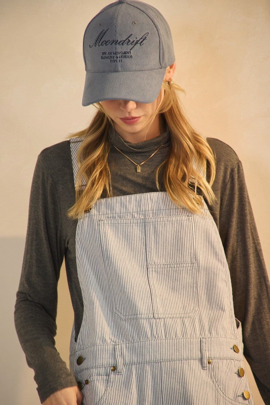 woman wearing a gray long sleeve top and blue striped overalls, accessorized with a blue cap and layered necklaces