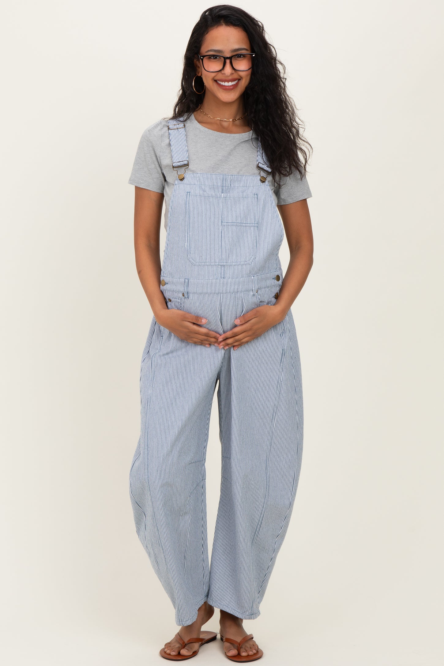 woman wearing a striped blue and white maternity overall with a gray t-shirt, holding her baby bump.