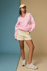woman wearing a pink v-neck sweater with white stripes and cream shorts, paired with a beige cap and sneakers