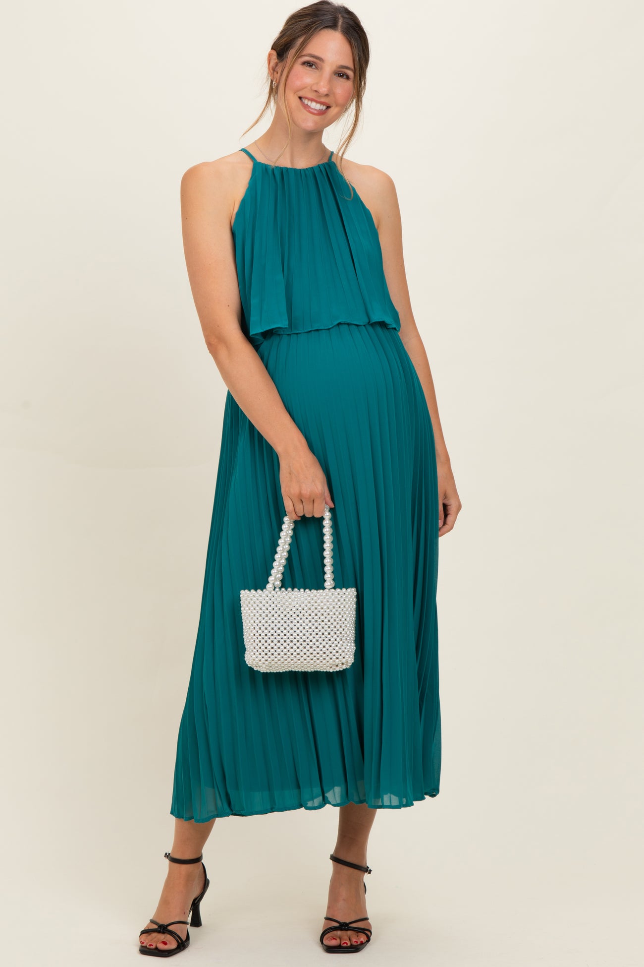woman wearing a teal pleated maternity dress with a flowing top and holding a white beaded handbag, showing her baby bump
