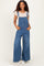 woman wearing a light pink short sleeve top and wide-leg denim overalls with pockets, smiling against a light background