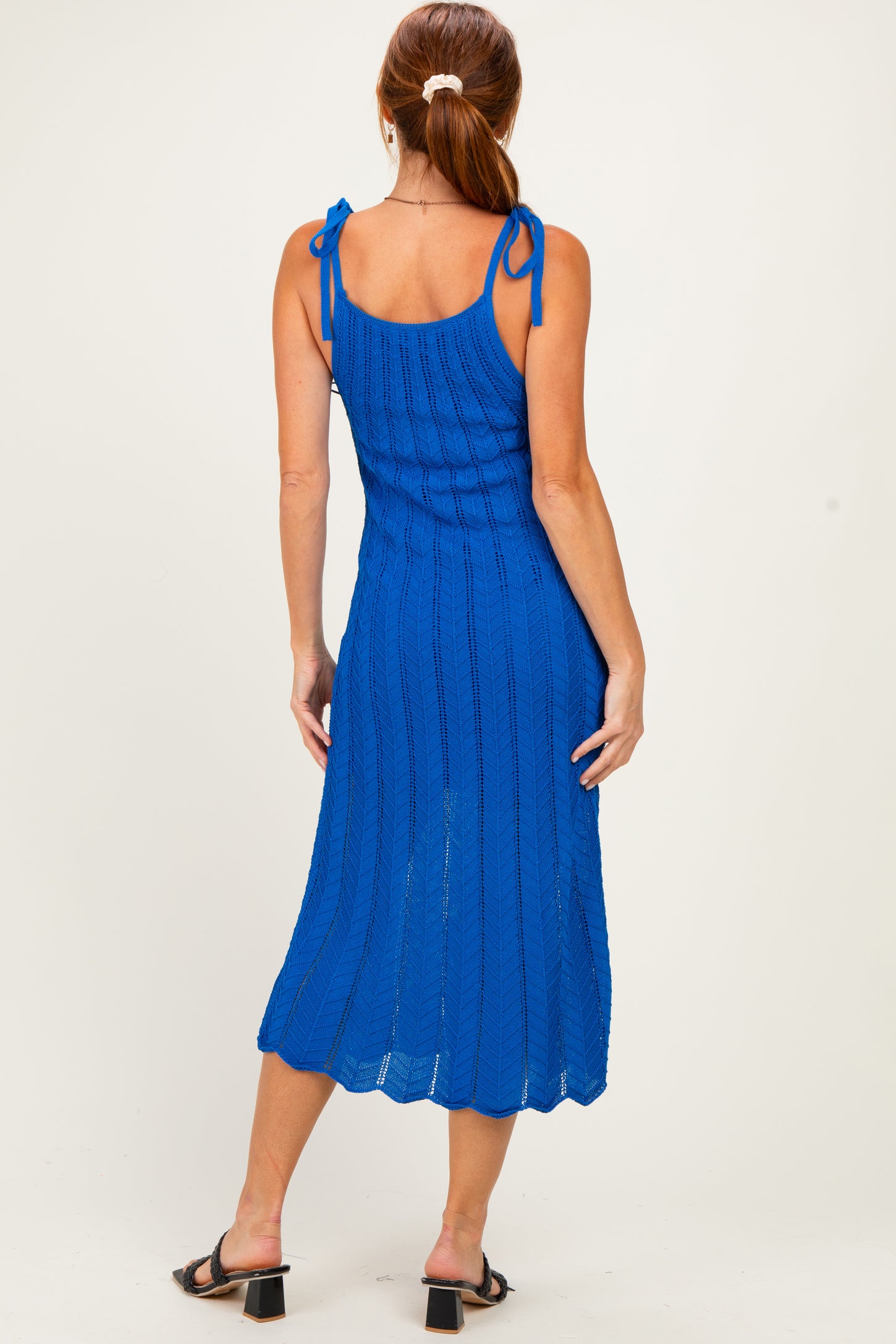 woman wearing a back view of a vibrant blue ribbed knit midi dress with adjustable tie straps and a scalloped hem