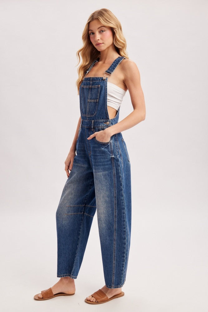 woman wearing a blue denim overall with wide legs and a white tank top underneath, standing with one hand in pocket