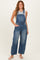 woman wearing a gray sleeveless top and blue denim overalls, standing with one hand in pocket