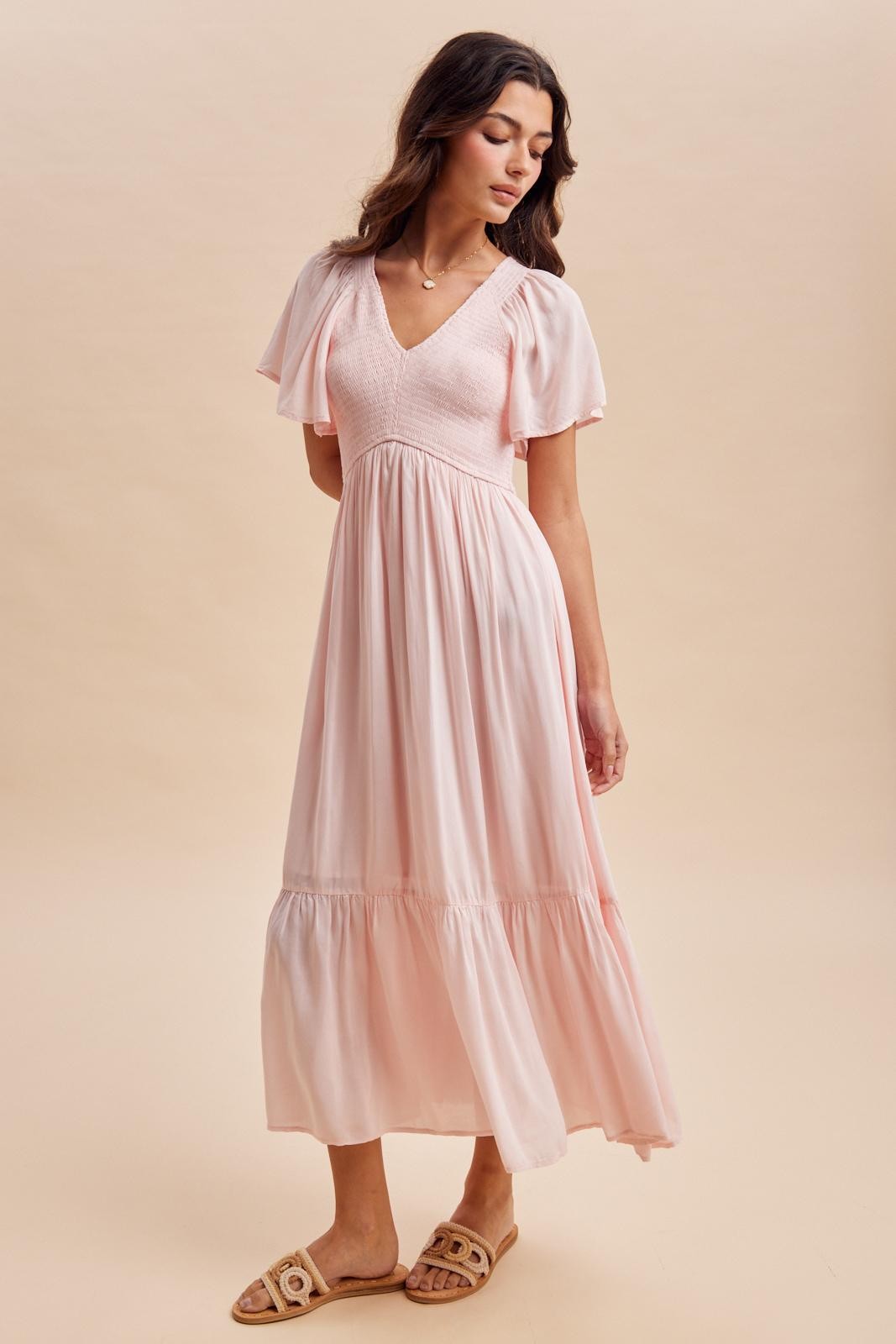 woman wearing a soft pink v-neck maxi dress with short flutter sleeves and a tiered skirt, styled with flat sandals