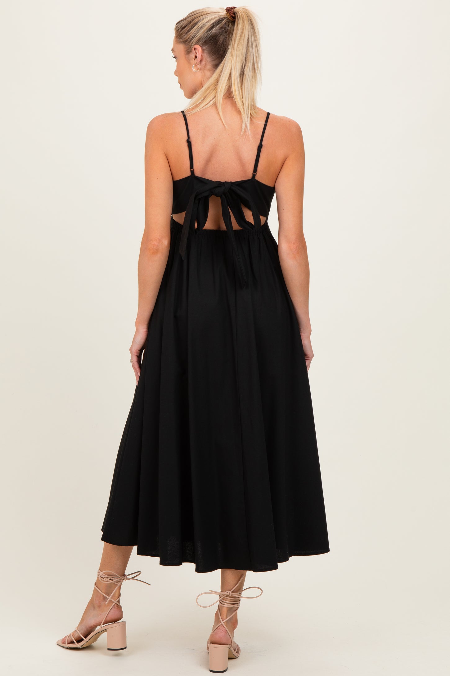 woman wearing a black maternity dress with open back and tie details, showing a flowing skirt and strappy sandals